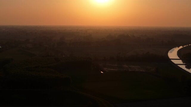 Po Valley in Lombardy Italy with agricultural fields and river corridor under low sun with haze and atmospheric scattering at sunset over flat alluvial plain, aerial shot