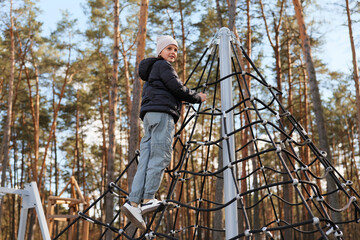 A child is climbing a netted structure at a playground located in a forest. The trees surround the play area under a clear blue sky © sementsova321