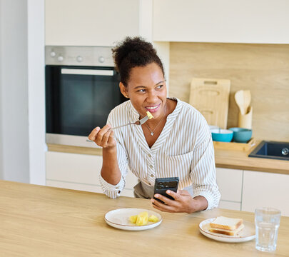 Portrait of a young woman or businesswoman gettyng ready for work preparing and eating breakfast fruit using a smartphone phone standing in kitchen home