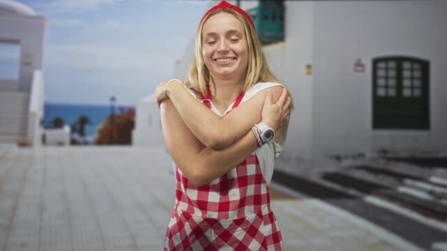 Woman hugging her arms, cook in red gingham apron and bandana on a seaside street with white buildings and stairs; self care contentment.