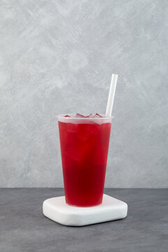 Iced red berry tea in plastic cup with drinking straw on white coaster. Cold hibiscus tea or fruit punch served in takeaway cup. Refreshing drink with ice cubes.