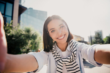 Fototapeta premium Young woman smiling and taking a selfie outdoors in sunny urban street, casual striped scarf, natural light portrait capturing happy lifestyle and travel vibes