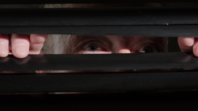 Woman opens horizontal blinds on window and eyes peer through hole into street with searching gaze close-up