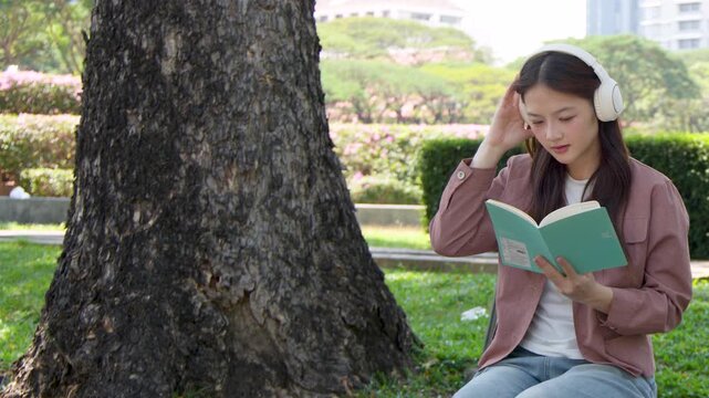 Young Asian Woman Reading Book and Wearing Headphones in a Sunny Outdoor Park
