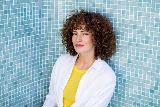 Confident model with curly hair leaning against blue pool tiles