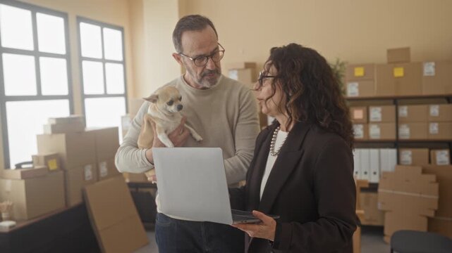 Man holding small dog while middle aged woman holds laptop and points to screen amid stacked parcels and boxes in a small business building; collaboration.