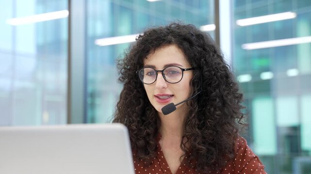 Female corporate operator in headset working in customer support service on helpline telesale talking using laptop sitting at workplace in business office. Agent of call center advises client Close up