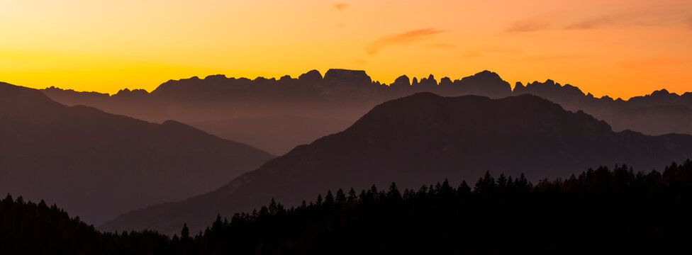 Mountain silhouettes and forest line against a vibrant sunset sky, an alpine nature tourism destination with copy space. Brenta Dolomites from Paganella plateau, Trentino, Italy.