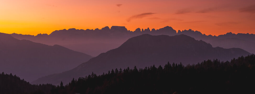 Layered mountain range and forest silhouettes against a vibrant orange sunset sky, showcasing a tranquil alpine travel destination. Brenta Dolomites from Paganella plateau, Trentino, Italy.