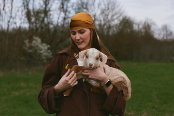 Naklejka premium Cheerful woman outdoors, smiling while holding a goat kid, gentle interaction, warm clothing, natural setting, soft light, close bond with animal, rustic lifestyle moment, countryside atmosphere
