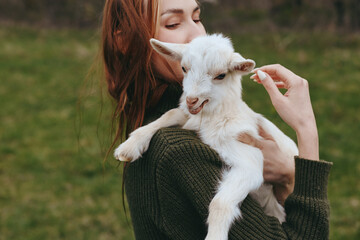Naklejka premium Goat, kid, woman outdoors, tender moment as she holds a gentle goat kid, sunlit grass, casual sweater, natural colors, candid lifestyle scene, outdoor companionship