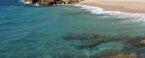 Obraz premium Clear turquoise ocean waves wash onto a sandy beach. Rocky formations border the water and shore. Sunlit water reveals underwater rocks and sand.