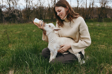 Naklejka premium Cheerful woman outdoors, feeding a friendly goat kid, warm sweater, soft grass, sunlit moment, relaxed posture, gentle interaction, natural colors, rural landscape
