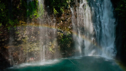 Naklejka premium Vibrant rainbow over a majestic tropical waterfall, a serene natural cascade flowing into a clear blue pool, lush green foliage framing the powerful water stream
