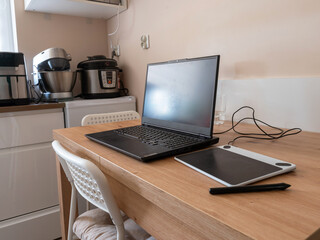 Fototapeta premium Laptop and Graphics Tablet on Wooden Kitchen Table for Remote Work and Digital Art. A black laptop and pen tablet sit on a wooden table in a kitchen environment.