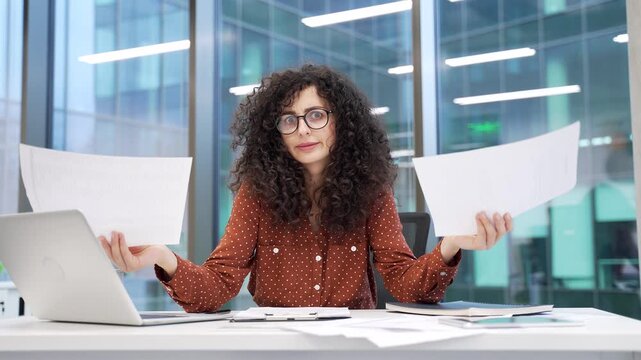 Disappointed businesswoman having difficulty with paperwork while sitting at workplace in a business office looking at camera. Puzzled frustrated female checks documents and cannot understand problem