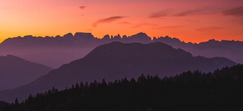 Layered mountain silhouettes and forest against a vibrant sunset sky, perfect for nature tourism and destination exploration. Brenta Dolomites from Paganella plateau, Trentino, Italy.