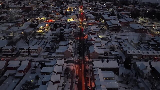 Aerial descent over Columbia, Pennsylvania during soft snowfall at night, with glowing orange streetlights, snow-covered rooftops, historic row houses and lit Main Street corridor. Flyover shot.
