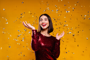 Naklejka premium Happy woman celebrating with confetti in red sequin dress on yellow background, festive party portrait expressing joy and glamour