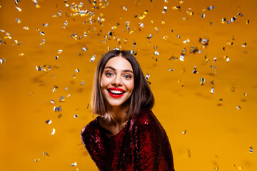Naklejka premium Smiling woman in red sequin dress celebrating with silver confetti against a vibrant yellow background, joyful glamorous party portrait