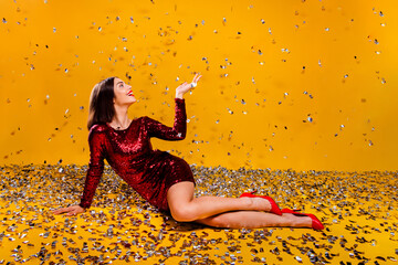 Naklejka premium Woman in red sequin dress lying among confetti on yellow background celebrating a festive party with glamorous heels and joyful expression