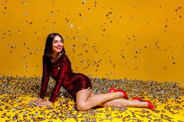 Naklejka premium Smiling woman in red sequin dress and high heels posing with falling confetti on yellow background for festive party celebration and glamorous studio portrait