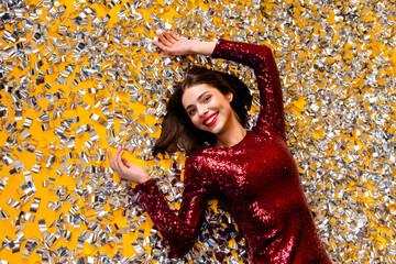 Naklejka premium Smiling woman in a red sequin dress lying in silver confetti for a festive party celebration and holiday glamour photoshoot