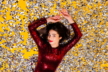 Naklejka premium Young woman in red sequin dress lying in silver confetti on yellow background celebrating party glamour and festive mood
