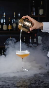 Bartender carefully pouring a cocktail with smoke effect into a glass, showcasing mixology and craft drinks in a bar setting.