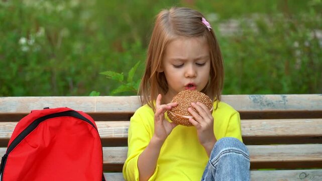 School child sits in school yard and eats hamburger. Student girl has breakfast outdoors. Education primary school preschool. Vertical shot