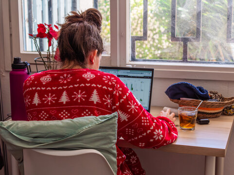 Mujer joven de espaldas teletrabajando con port&aacute;til en casa con un jersey navide&ntilde;o rojo