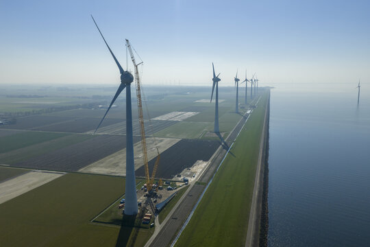 Aerial view of wind turbines standing in a row along a coastal dike with a construction crane and flat green fields under a clear blue sky.