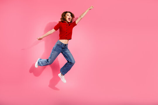 Joyful young woman jumps in the air against pink background wearing red top and blue jeans lifestyle fashion