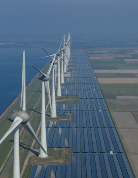 Aerial view of a row of modern wind turbines and vast solar panel arrays situated along a coastal dike under a clear blue sky.