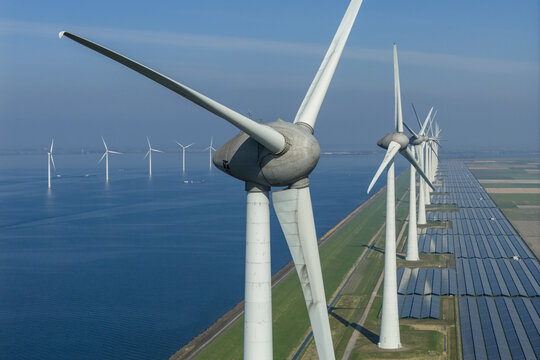 Aerial view of a large-scale renewable energy park featuring rows of wind turbines and solar panels along a coastal dike under a clear blue sky.