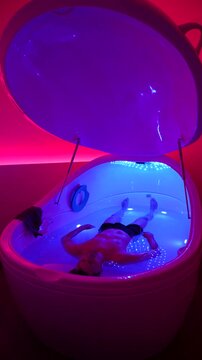 Man floats in Epsom salt water inside a floatation therapy pod, neon wellness scene, static vertical shot.