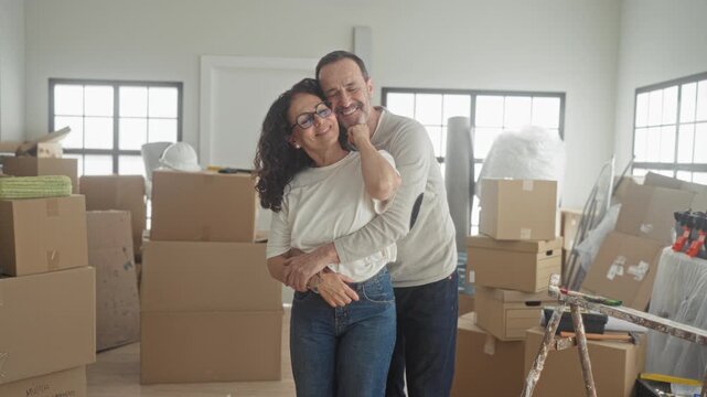 Man hugs woman from behind, hands around waist, smiling amid moving boxes and ladder in living room; moving day contentment.