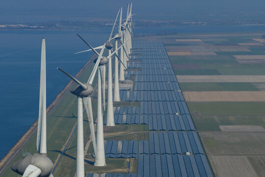 Aerial view of a long row of modern wind turbines and an extensive solar panel farm located along a coastal dike next to cultivated agricultural fields and a calm body of water.