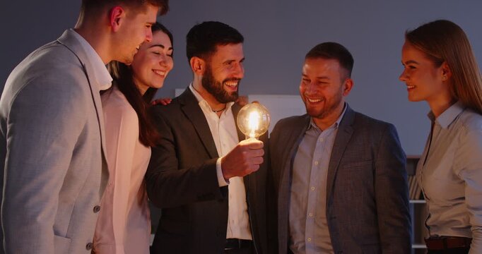 Team of successful women and men coworkers meeting together for solving problem, found nice idea, holding light bulb and standing in half circle. Team work and brainstorming concept