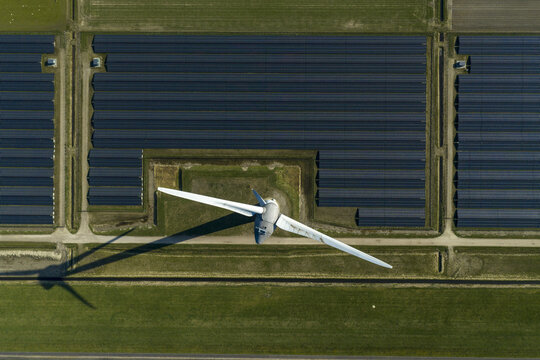 Aerial view of a large wind turbine and rows of solar panels creating geometric patterns on green fields in Creil, Flevoland, Netherlands.