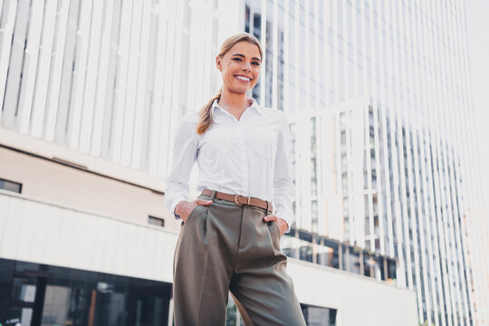 Confident young business woman smiling in front of modern city office building wearing white shirt and tailored trousers, professional urban portrait conveying success and optimism