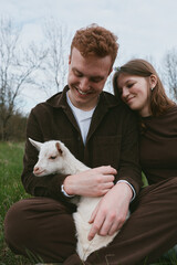 Naklejka premium A couple shares a gentle moment with a cheerful goat kid in a grassy meadow, embracing outdoors, showing rural lifestyle and affectionate bond, serene skies, casual clothing, warm interaction,