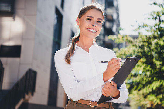 Smiling businesswoman outdoors holding clipboard and pen conducting inspection or real estate survey, confident professional in white shirt and trousers presenting clipboard notes