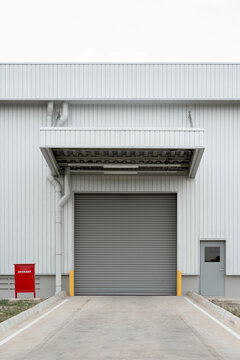 Industrial warehouse loading dock with closed roller door, canopy, grey personnel door, yellow bollards, and a red fire hydrant box.