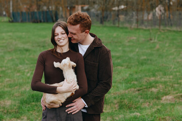 Naklejka premium A joyful couple shares a moment with a small goat kid in an open grassy park, embracing and smiling. A warm lifestyle scene that captures affection, companionship, and simple countryside moments.