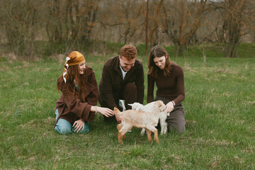 Naklejka premium Goat kid animal family outdoor field nature interaction three people gently guide a playful goat kid in a grassy field, capturing a warm, candid moment of care and connection
