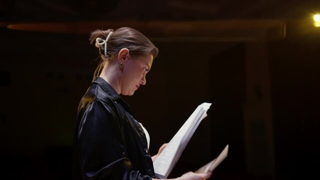 Young actor rehearsing dramatic script under stage lights in dark empty theater, practicing lines with focused concentration during theatrical performance preparation