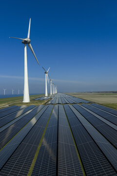 Aerial view of a vast solar panel farm and a row of tall wind turbines along the coast and green fields under a clear blue sky Netherlands.