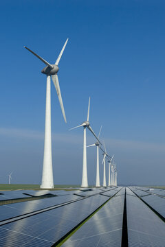 Aerial view of a row of tall wind turbines integrated with a large-scale solar panel array under a bright blue sky in the Netherlands.