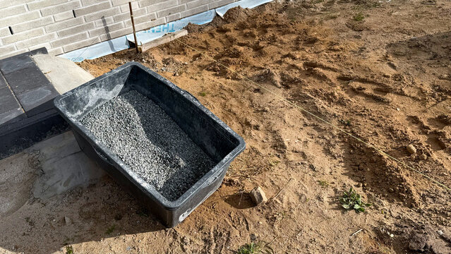 Black plastic container filled with gravel placed on sandy ground near a building foundation, with construction tools and materials visible in the background
